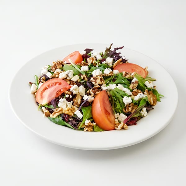 Fresh green salad with walnuts, feta cheese, dried cranberries, and tomato slices on a white plate.